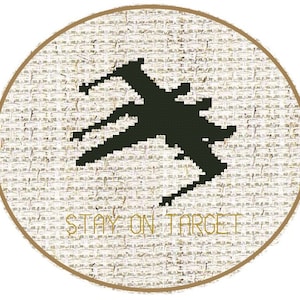 May include: A round, beige fabric design with a dark green pixelated spaceship silhouette. The text "STAY ON TARGET" is in gold lettering below the spaceship. The design is framed by a thin, gold-colored border.