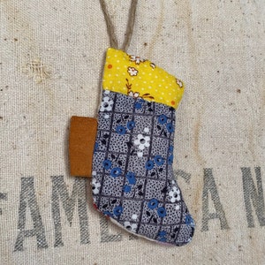 May include: A small, handmade stocking ornament with a yellow, white, and blue floral pattern. The stocking is made of fabric and has a brown leather patch on the side. It is hanging from a twine string.