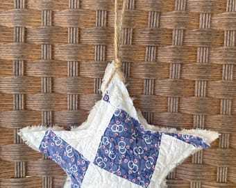 Puede incluir: Un adorno de estrella de patchwork azul y blanco con borde deshilachado. La estrella cuelga de una cuerda de yute marrón.