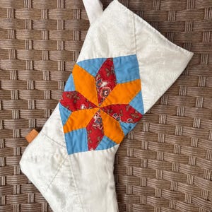 May include: A white quilted Christmas stocking with a patchwork design featuring orange, red, and blue fabric. The stocking has a loop at the top for hanging.
