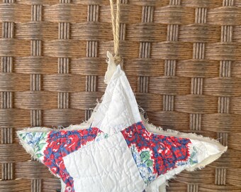 Puede incluir: Un adorno de estrella de retazos blanco, rojo y azul con un borde deshilachado. La estrella cuelga de una cuerda de yute marrón.