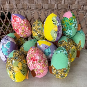 May include: A collection of handmade fabric Easter eggs in various colours and patterns, including purple, green, yellow, pink, and blue. The eggs are made from patchwork fabric and have a rustic, vintage aesthetic.