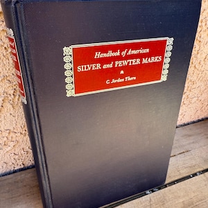 May include: A black hardback book titled "Handbook of American SILVER and PEWTER MARKS" by C. Jordan Thorn. The title is in gold lettering on a red background with a gold decorative border.