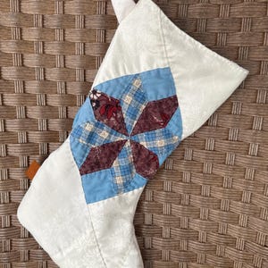 May include: A quilted Christmas stocking with a starburst design. The stocking is primarily white with a floral pattern, and features a blue and burgundy star in the center. A white loop is attached for hanging.