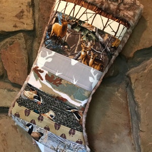 May include: A Christmas stocking with a patchwork design featuring wildlife and camouflage patterns. The stocking includes images of deer, ducks, and footprints, with a faux fur trim at the top. The color palette includes browns, greens, and tans.