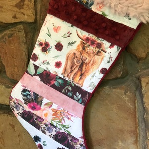 May include: A Christmas stocking with a patchwork design. The top is lined with faux fur and a burgundy fabric. The main body features a watercolor image of a Highland cow and calf, with floral and striped patterns.