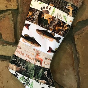 May include: A Christmas stocking with a variety of outdoor-themed patterns. The stocking features images of fish, deer, ducks, and paw prints. The text "Little Man" is printed on the stocking. The top of the stocking is trimmed with faux fur.
