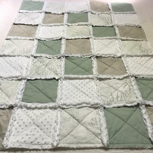May include: A handmade patchwork quilt featuring a variety of fabrics in shades of green, white, and tan. The quilt is composed of square blocks with different patterns, including solid colors, dotted textures, and botanical prints. The edges are finished with a ruffled trim.