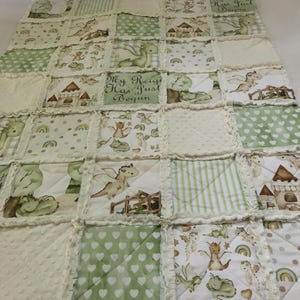 May include: A patchwork quilt featuring a repeating pattern of green dragons, castles, and rainbows on a white background. The quilt has a ruffled edge and the text "My Reign Has Just Begun" is visible on some squares. The quilt is made of soft fabric.