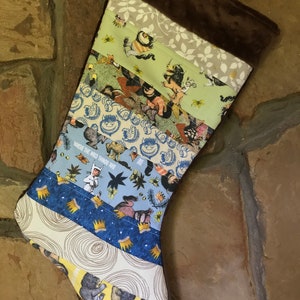 May include: A handmade Christmas stocking with a patchwork design. The stocking features various fabrics with illustrations from "Where the Wild Things Are." The top is a dark brown plush material. The stocking is designed to be hung by a fireplace.