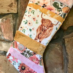 May include: A Christmas stocking featuring a patchwork design with floral patterns in shades of pink, orange, and green, a central Highland cow illustration, a mustard yellow fabric band, a tan faux fur section, and a soft pink faux fur cuff.