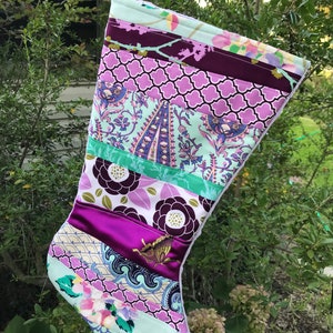 May include: A handmade Christmas stocking with a patchwork design. The stocking features various patterned fabrics in shades of purple, green, and white. A decorative butterfly is sewn onto a purple satin stripe. A purple ribbon is attached for hanging.