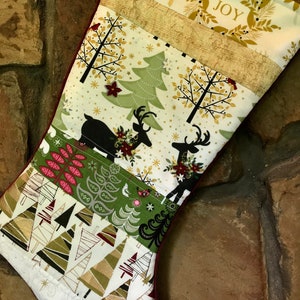 May include: A Christmas stocking with a festive design. The top features a gold reindeer and the word "JOY" in a wreath. The body has various patterns of trees, reindeer, and floral accents in green, gold, and red. The stocking is trimmed with a burgundy fabric.