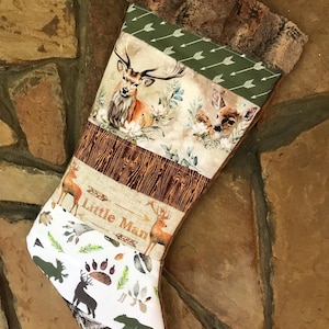 May include: A Christmas stocking featuring a patchwork design with a faux fur cuff. The fabric panels display illustrations of deer, wood grain patterns, arrow motifs, and forest animal silhouettes. The text Little Man is printed on the fabric.