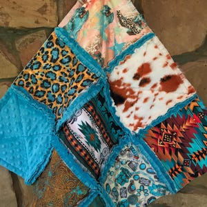 May include: A patchwork quilt with a variety of patterns and textures. The quilt features squares of turquoise, leopard print, cowhide, and Southwestern designs. The edges are trimmed with a ruffled turquoise fabric. The quilt is hanging against a stone wall.