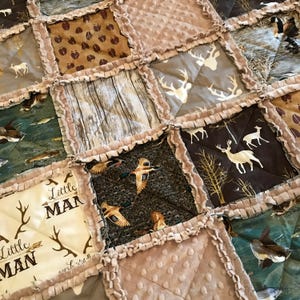 May include: A patchwork quilt with various hunting-themed patterns. The quilt features squares with deer, ducks, and wood grain designs. The text "Little Man" is printed on a cream-colored square. The edges are trimmed with a ruffled border.