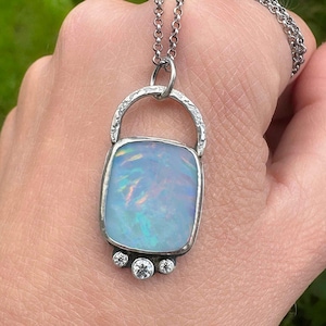 Könnte beinhalten: Silberne Anhängerkette mit einem großen, quadratischen Opal mit schillernden Blau- und Grüntönen. Der Anhänger ist mit drei kleinen, klaren Edelsteinen verziert und hängt an einer filigranen Kette.