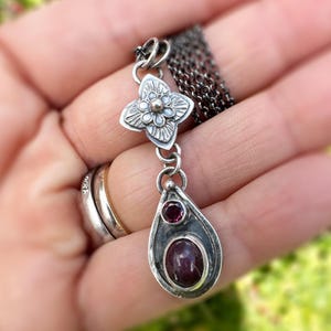 Può includere: Una collana con pendente in argento con un motivo floreale e un pendente a forma di goccia con due pietre preziose rosse. La collana ha una catena scura. I dettagli intricati del gioiello sono visibili, tenuti in una mano.