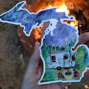 May include: A white sticker shaped like the state of Michigan with a watercolor sky with stars and a crescent moon. The sticker has a small camper van parked in a forest with two people sitting by a campfire.