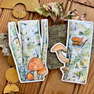 May include: Two watercolor illustrations of birch trees with mushrooms growing on them. The first illustration features a cluster of red and white mushrooms with white spots. The second illustration features a single orange mushroom with a white stem.