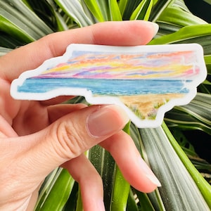 May include: A sticker of the state of North Carolina with a watercolor painting of a sunset over the ocean. The sticker is white with a blue, pink, orange, and yellow sunset over a blue ocean with a sandy beach.