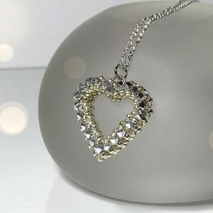 May include: A silver chain necklace with a silver and gold beaded heart pendant. The pendant has a small open space in the center of the heart. The necklace is displayed on a clear glass surface.