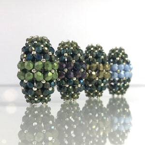 Op de afbeelding: Vier kralenbollen in tinten van groen, paars, bruin en blauw. De kralen zijn in een rij op een wit oppervlak gerangschikt.