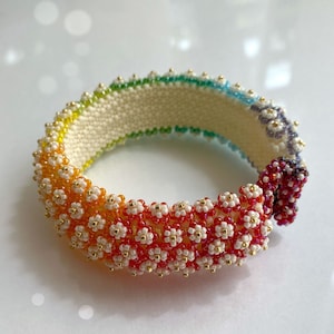 Op de afbeelding: Een regenboogkleurige kralenarmband met witte bloemvormige kralen en gouden accenten. De armband is gemaakt met verschillende kleuren, waaronder rood, oranje, geel, groen, blauw en paars.