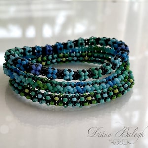 May include: A set of four beaded bracelets in shades of blue, green, and black. The bracelets are stacked on top of each other, creating a layered effect.