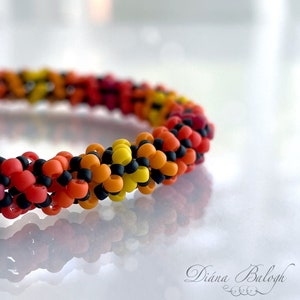May include: A close-up of a colorful beaded bracelet with a gradient of red, orange, yellow, and black beads. The bracelet is made with small, round beads and is coiled into a spiral shape.