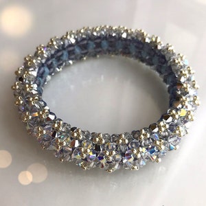 Puede incluir: Una pulsera de cuentas circular con un interior azul claro y un exterior brillante similar al cristal. La pulsera presenta pequeños detalles dorados. La imagen está bien iluminada, mostrando los intrincados detalles de la joyería.