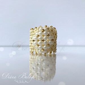 May include: A white and gold beaded ring with a geometric design. The ring is made with small, round beads and is a statement piece.