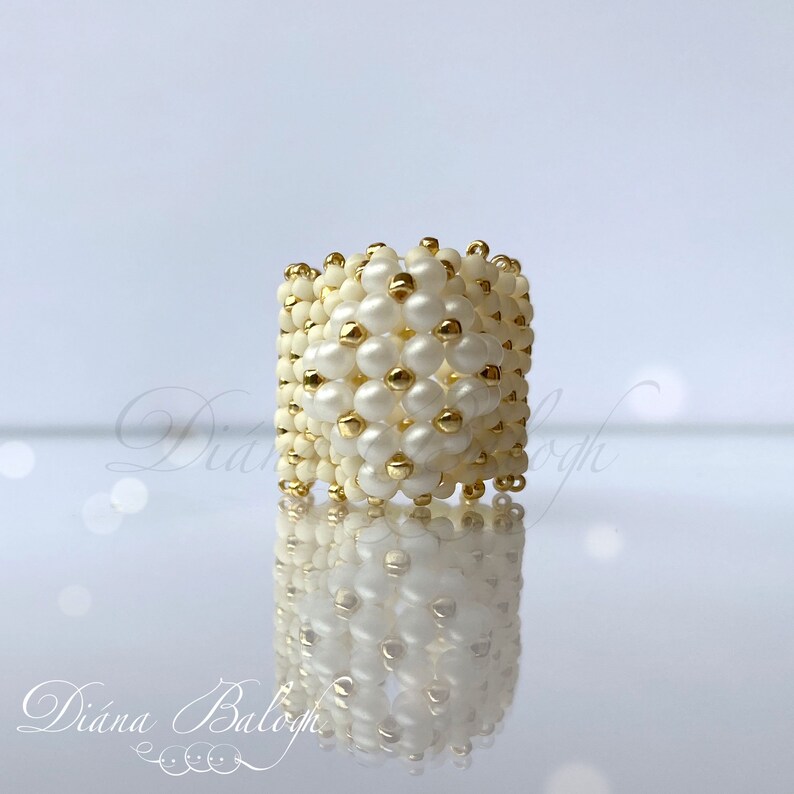 May include: A white and gold beaded ring with a geometric design. The ring is made with small, round beads and is a statement piece.