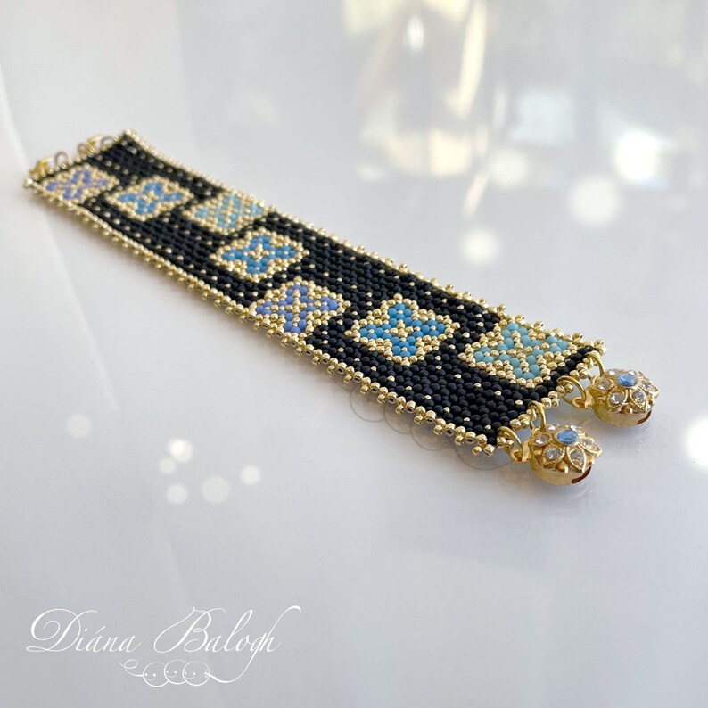 Puede incluir: Un brazalete de cuentas negras con detalles dorados y azules. El brazalete presenta un patr&oacute;n geom&eacute;trico y tiene cierres dorados con piedras transparentes.