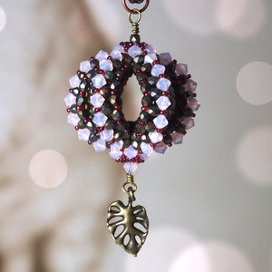 May include: A beaded pendant with a spiral design in shades of purple, black, and brown. The pendant is attached to a bronze chain with a leaf-shaped charm.