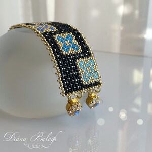 Puede incluir: Un brazalete de cuentas negras y doradas con detalles azules y turquesa. El brazalete tiene un patr&oacute;n geom&eacute;trico y cuenta con colgantes con cristales transparentes.