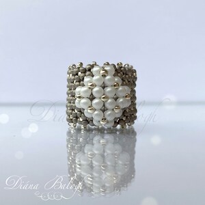May include: A silver and white beaded ring with a geometric design. The ring is made with small, round beads and has a square shape.