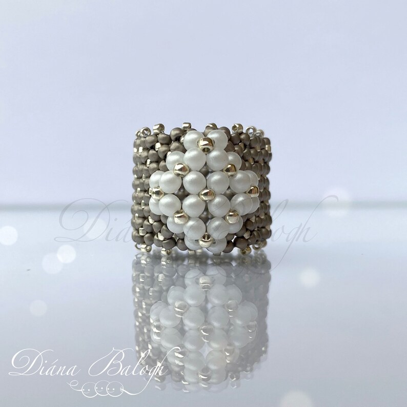 May include: A silver and white beaded ring with a geometric design. The ring is made with small, round beads and has a square shape.