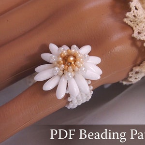 May include: A beaded ring with a white daisy flower design. The flower has white petals and a gold center, with small white beads around the petals. The ring is on a finger. Text at the bottom reads "PDF Beading Pattern".