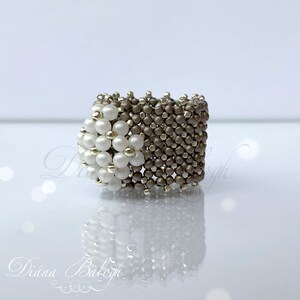 May include: A white and gray beaded ring with a flower design. The ring is made of small, round beads that are woven together in a intricate pattern.