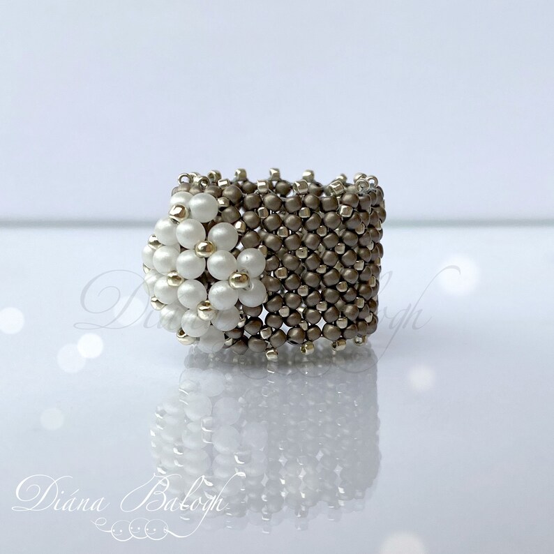 May include: A white and gray beaded ring with a flower design. The ring is made of small, round beads that are woven together in a intricate pattern.