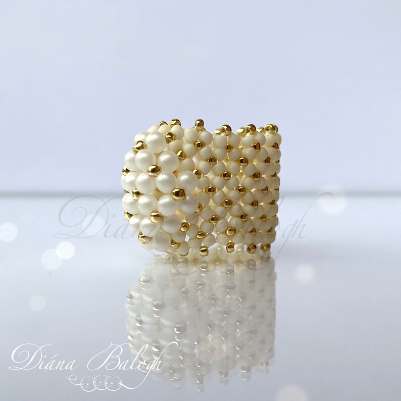 May include: A white and gold beaded ring. The ring is made of small, round beads that are arranged in a square pattern. The beads are white with gold accents.