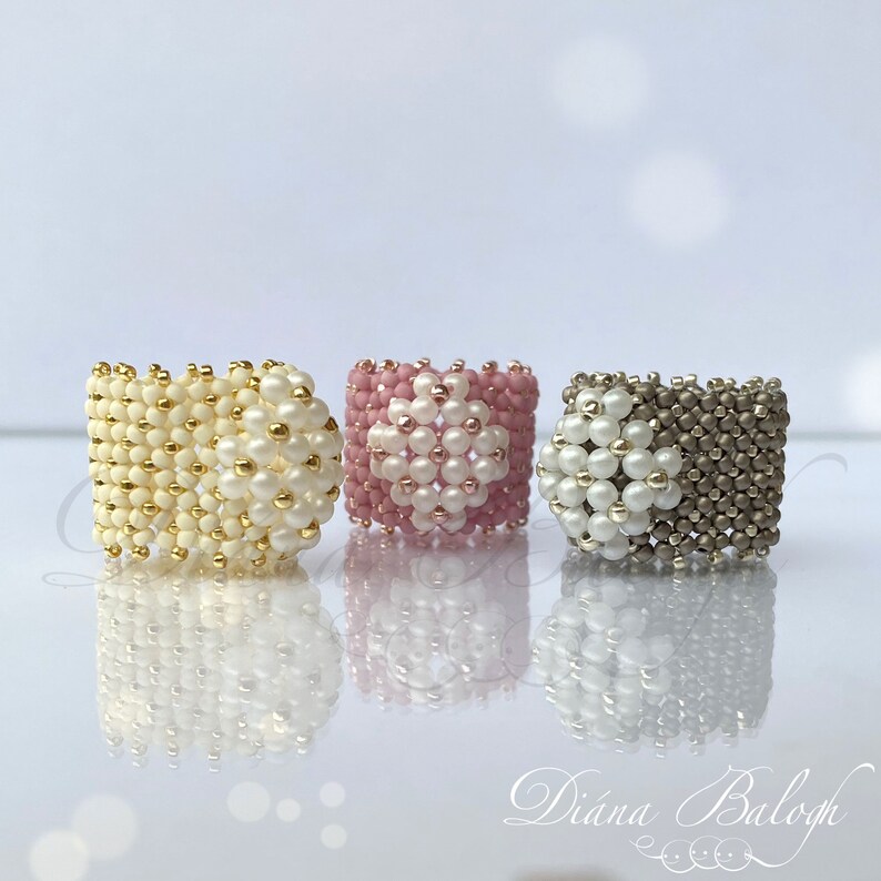 May include: Three beaded rings with a white, pink, and gray color scheme. Each ring features a white flower design in the center.
