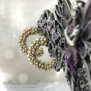 May include: Close-up of a pair of cream-colored beaded earrings with gold accents. The earrings are hanging from a silver fairy figurine with purple wings and flowers.