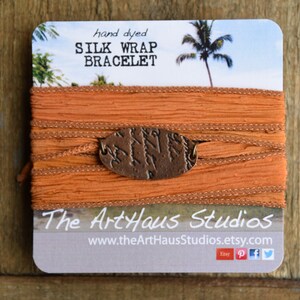 Puede incluir: Pulsera de envoltura de seda naranja teñida a mano con un dije de cobre. La pulsera está empaquetada en una tarjeta con el texto "hand dyed SILK WRAP BRACELET" y "The ArtHaus Studios www.theArtHausStudios.etsy.com".
