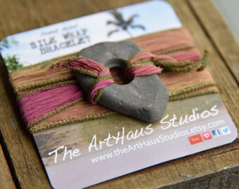 Könnte beinhalten: Ein grauer Stein mit Loch ist mit rosa und grünem Seidenband umwickelt und liegt auf einer Karte mit dem Text "Silk Wrap Bracelet". Die Karte zeigt auch den Text "The ArtHaus Studios" und Social-Media-Symbole.