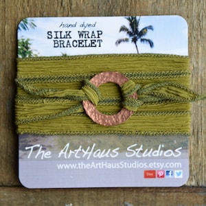 Pode incluir: Um bracelete de seda verde-oliva tingido à mão com um fecho de anel de cobre martelado. O bracelete é apresentado num cartão com o texto "hand dyed SILK WRAP BRACELET" e "The ArtHaus Studios www.theArtHausStudios.etsy.com".