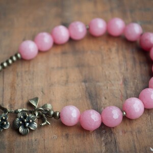 May include: A pink beaded bracelet with a bronze floral charm. The bracelet is made of round, faceted beads and has a delicate, feminine design.