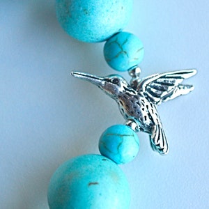 Op de afbeelding: Turquoise edelsteen kralen met een zilveren kolibrie bedel. De kralen zijn rond en hebben een gladde, gepolijste afwerking. De kolibrie bedel is gedetailleerd en heeft een delicaat ontwerp.