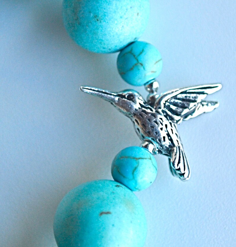 Op de afbeelding: Turquoise edelsteen kralen met een zilveren kolibrie bedel. De kralen zijn rond en hebben een gladde, gepolijste afwerking. De kolibrie bedel is gedetailleerd en heeft een delicaat ontwerp.